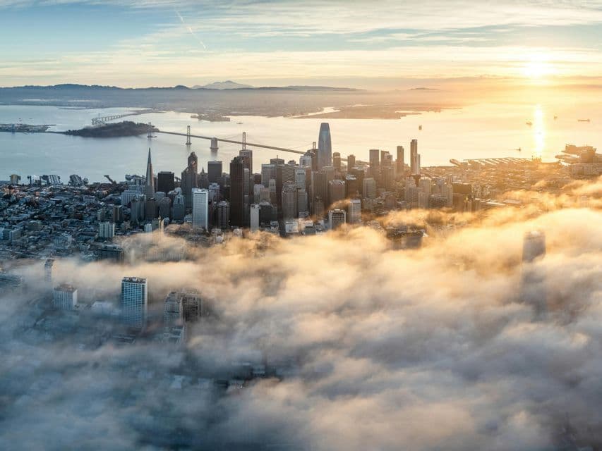 An aerial view of a city skyline with skyscrapers rising through a thick blanket of clouds at sunrise over a bay.