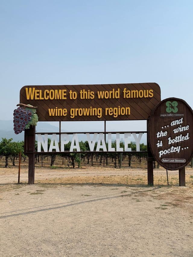 The official welcome sign for the Napa Valley wine growing region, with vineyards visible in the background under a hazy sky.