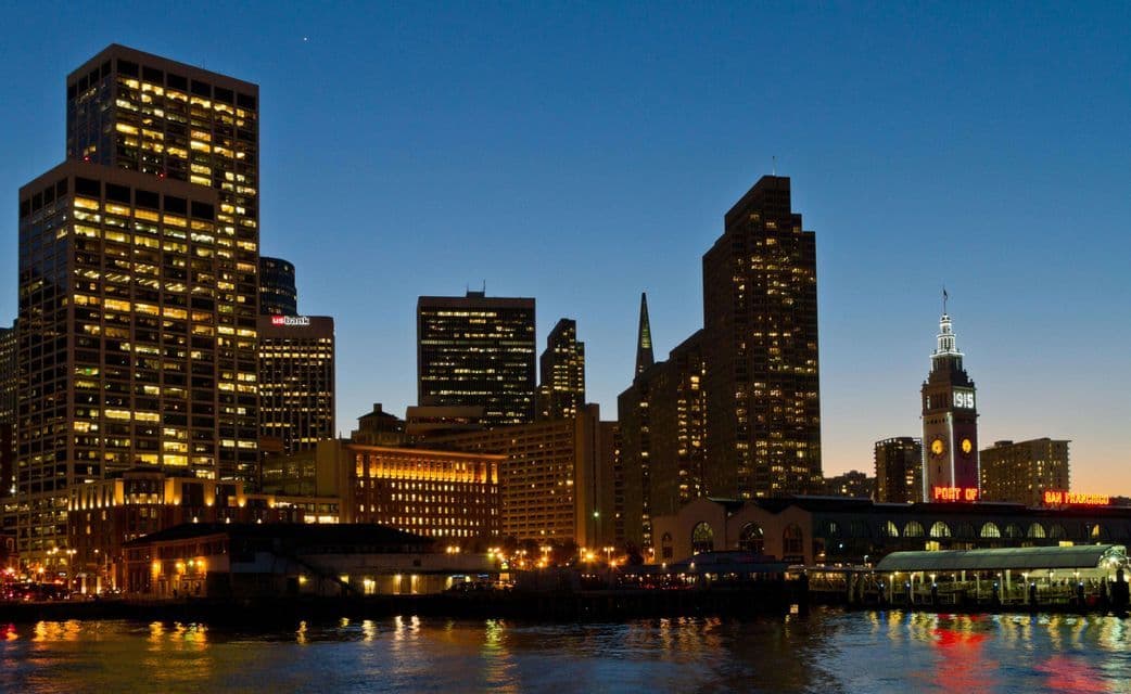 An illuminated city skyline with skyscrapers and a clock tower reflecting in the water at dusk.