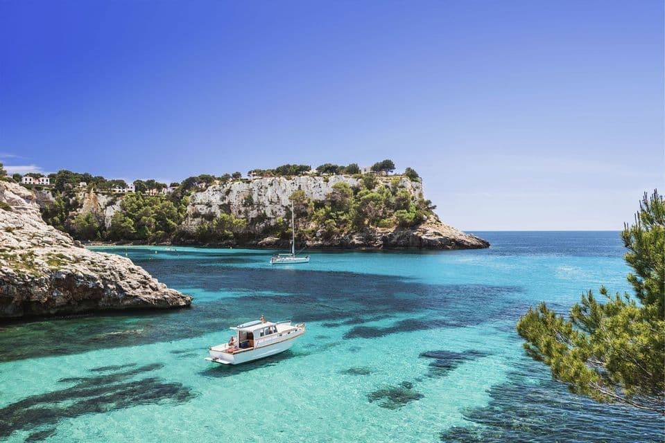 Barche galleggiano su acqua limpida e turchese in una caletta circondata da scogliere rocciose e alberate sotto un cielo azzurro brillante.