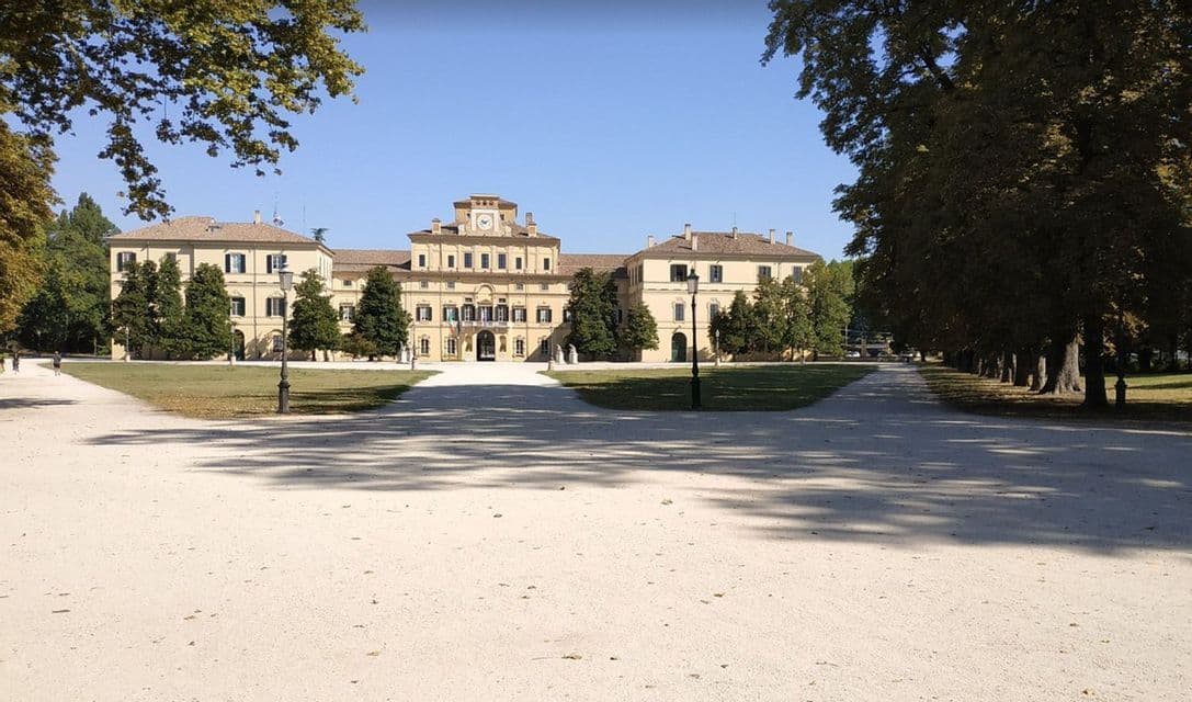 Un imponente edificio classico con una torre dell'orologio si erge dietro un prato verde e un ampio cortile di ghiaia, incorniciato da alberi.
