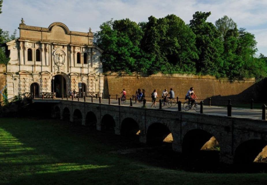 Un viaggio di gruppo WeRoad camminando e pedalando attraverso un ponte di pietra ad arco che conduce a una storica porta d'accesso e alla cinta muraria.
