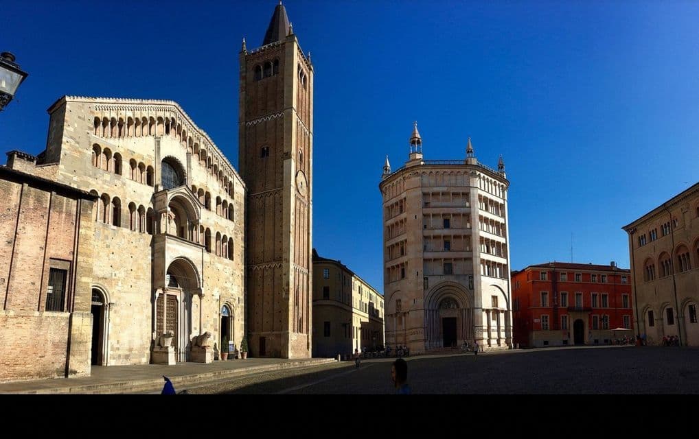 Una cattedrale storica in pietra, un campanile in mattoni e un battistero si ergono in una piazza assolata e aperta sotto un cielo azzurro e limpido.