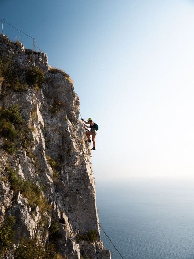 Una persona con il casco affronta una via ferrata su un'alta parete rocciosa a picco sull'oceano.