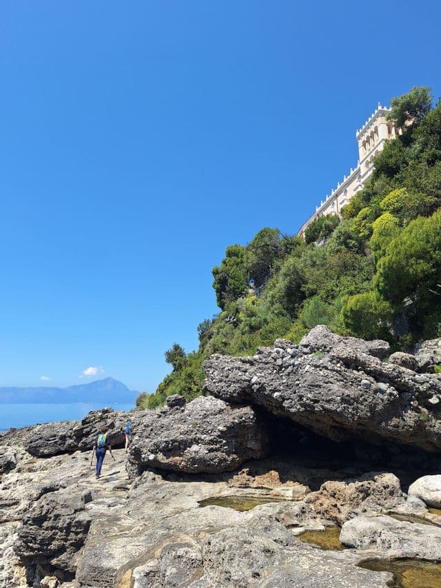 Un escursionista con uno zaino cammina lungo un sentiero costiero roccioso a picco sul mare, con un edificio su una scogliera verde in alto.