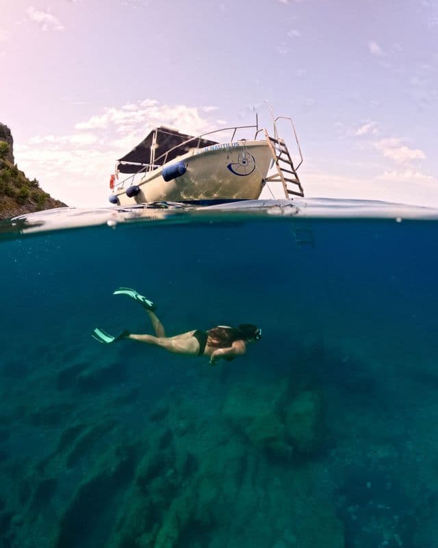 Foto split-shot di una persona che fa snorkeling nel mare cristallino, con una barca ancorata in superficie.