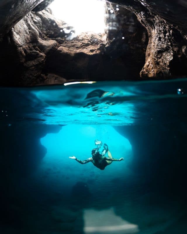 Immagine a vista divisa di una persona che nuota sott'acqua in una grotta marina baciata dal sole, con il soffitto roccioso visibile sopra il livello dell'acqua.