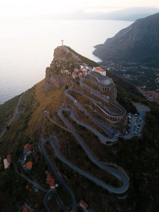 Veduta aerea di una strada tortuosa con tornanti che si inerpica su una ripida montagna costiera verso una statua in vetta.