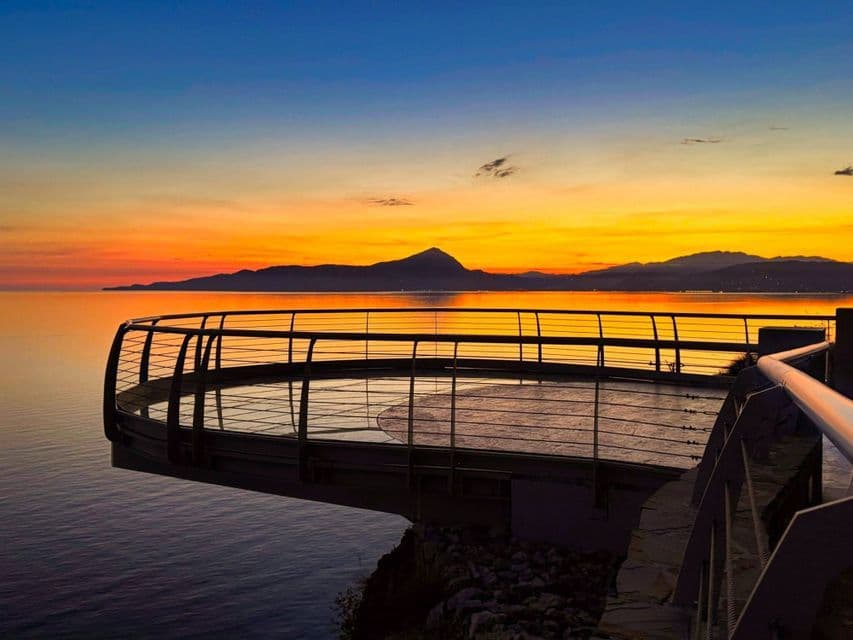 Una moderna piattaforma panoramica circolare si protende su un mare calmo, di fronte a montagne lontane sotto un cielo vibrante al tramonto.