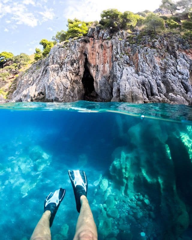 Veduta subacquea e di superficie di una persona che fa snorkeling vicino a una scogliera rocciosa con ingresso a una grotta.