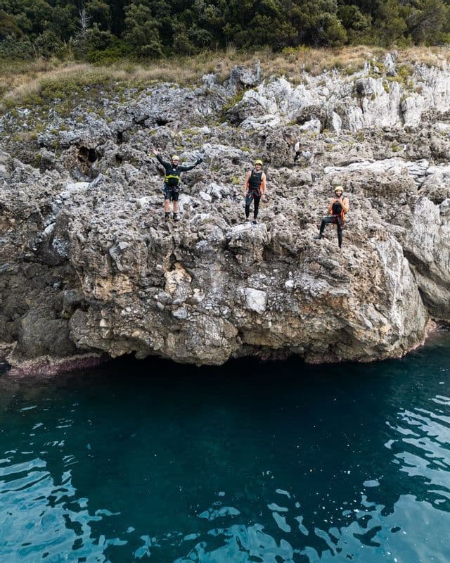 Un gruppo WeRoad di tre persone, con caschi e imbracature, si trova su una scogliera rocciosa a picco su acque blu profonde.