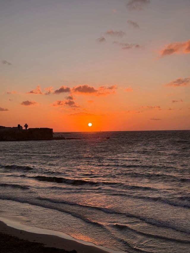 Un sole arancione cala sull'oceano, con onde che si infrangono su una spiaggia sabbiosa e una fortificazione stagliata sulla costa.