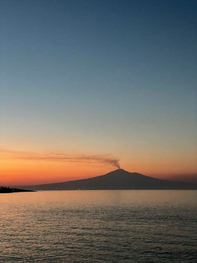 Un vulcano in silhouette rilascia una colonna di fumo sull'acqua sotto un cielo sfumato di arancio e blu al tramonto.