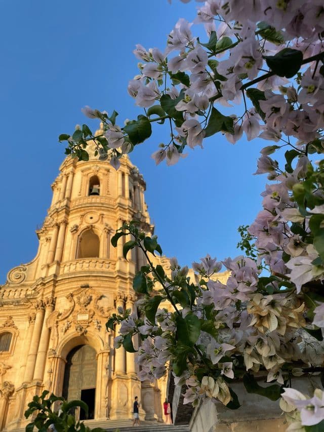La facciata ornata di una cattedrale color sabbia, incorniciata da fiori di bouganville rosa chiaro contro un cielo azzurro limpido.