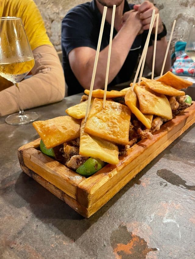 Un tagliere di legno con fritti, spiedini e spicchi di lime su un tavolo, accanto a un bicchiere di vino bianco.