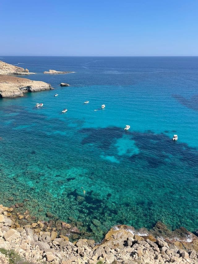 Vista dall'alto di una costa rocciosa con diverse piccole imbarcazioni che galleggiano su acque turchesi e cristalline sotto un cielo azzurro.