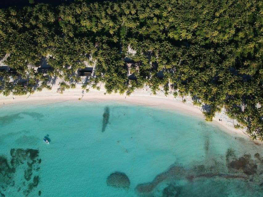Panoramica aerea di una spiaggia di sabbia bianca orlata di palme, accanto a limpide acque oceaniche turchesi con una piccola barca.