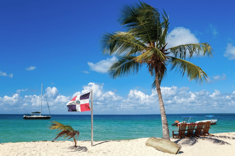 Una bandiera della Repubblica Dominicana si erge su una spiaggia di sabbia bianca accanto a una palma, con barche sul mare turchese sotto un cielo azzurro.