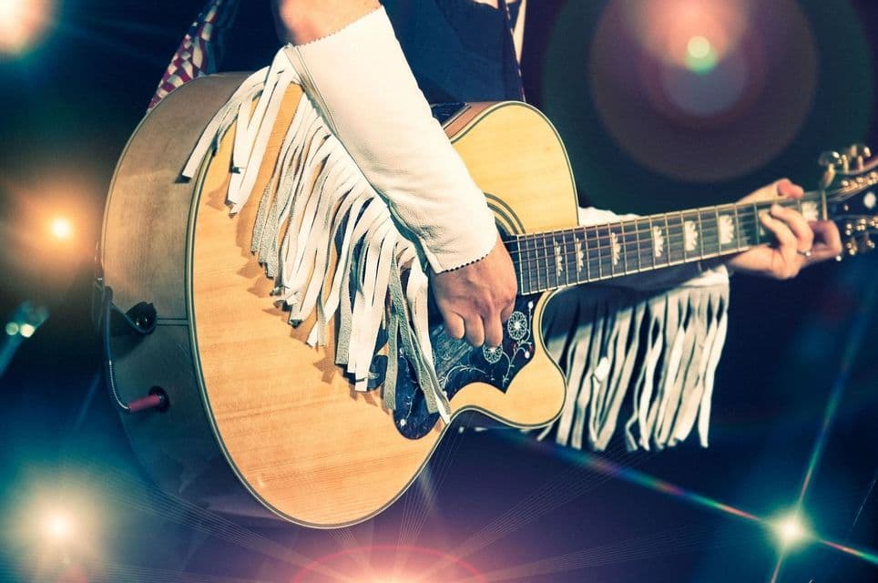 A close-up of a musician playing an acoustic guitar with a white fringed strap on stage, surrounded by lens flare.