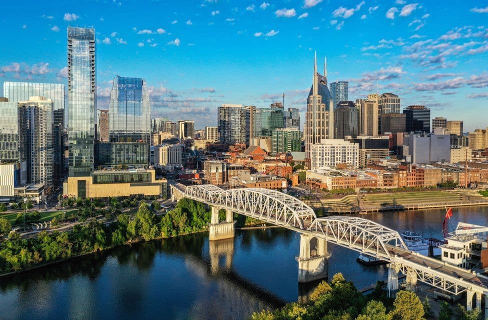An aerial view of a white truss bridge over a river, with a modern city skyline of skyscrapers in the background under a blue sky.