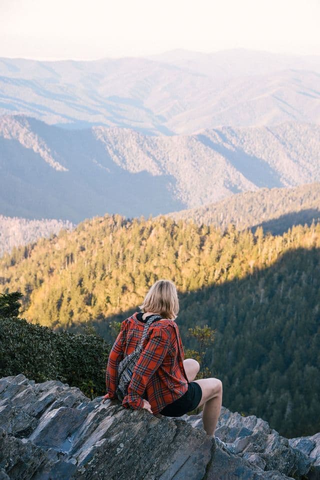 A person wearing a red plaid shirt sits on a rocky ledge, looking out over a vast, forest-covered mountain range.