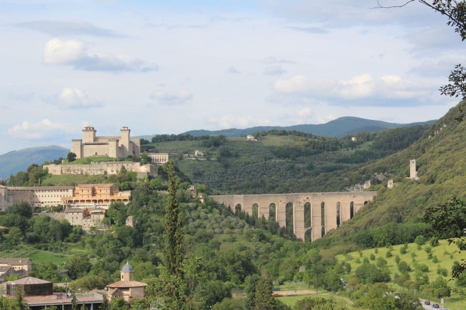 Un castello in pietra e un acquedotto dominano una città su una collina verde lussureggiante con montagne sullo sfondo sotto un cielo nuvoloso.