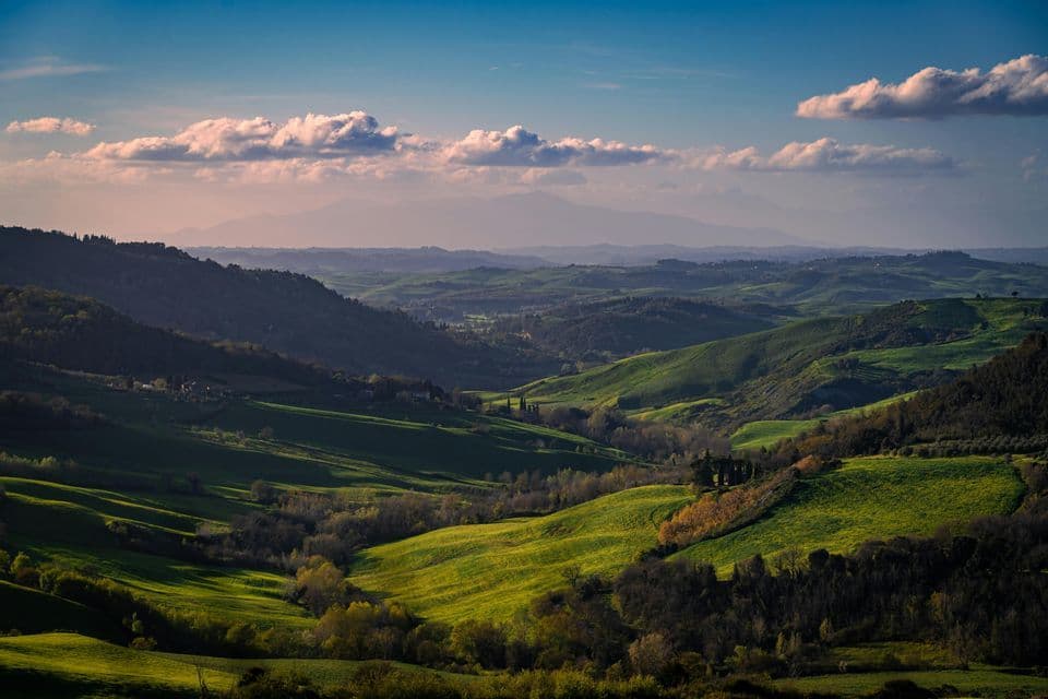 Un vasto paesaggio di dolci colline e valli verdi, illuminate da sole e ombra, con montagne in lontananza sotto un cielo blu.