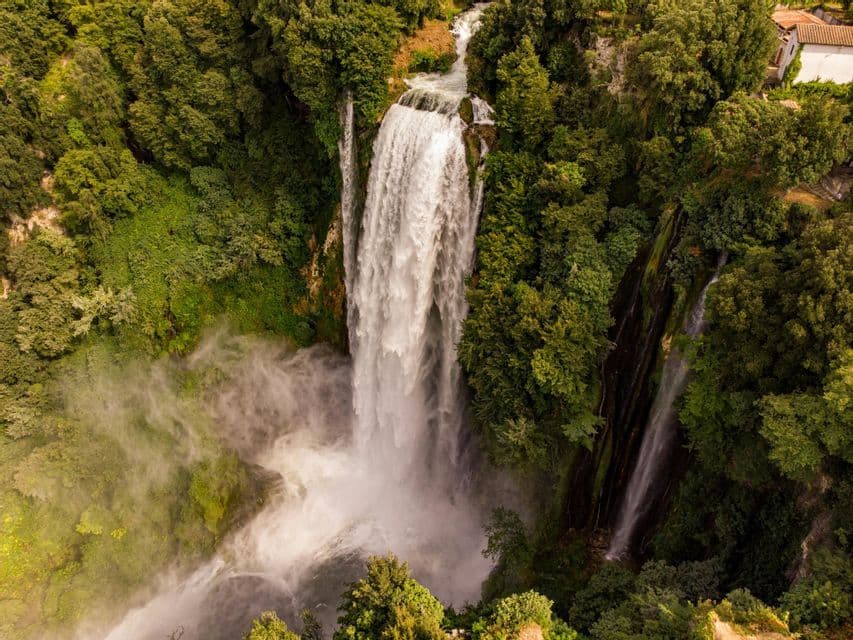 Una vista aerea di una grande cascata che scende da una scogliera rocciosa, circondata da una fitta foresta verde, creando un pennacchio di nebbia.