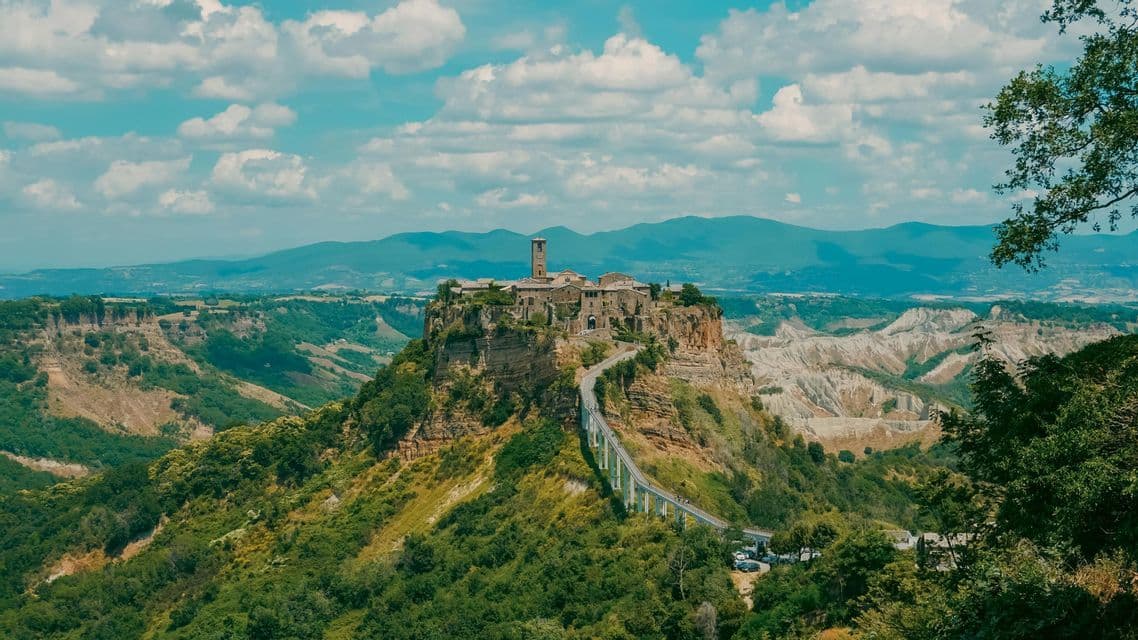 Un borgo storico con un campanile si erge su una scogliera rocciosa, collegato da un lungo ponte su una valle di verdi colline e calanchi.