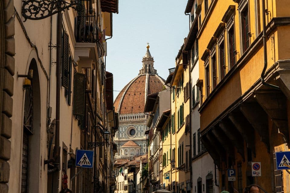 Una grande cupola di cattedrale con tegole rosse è visibile al termine di una strada stretta, fiancheggiata da alti edifici storici.