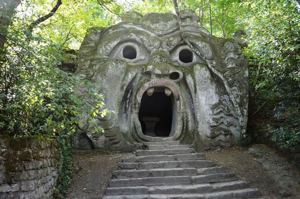 Una grande scultura in pietra ricoperta di muschio, raffigurante un volto con la bocca spalancata che funge da ingresso, situata in una foresta con scalini in pietra che vi conducono.