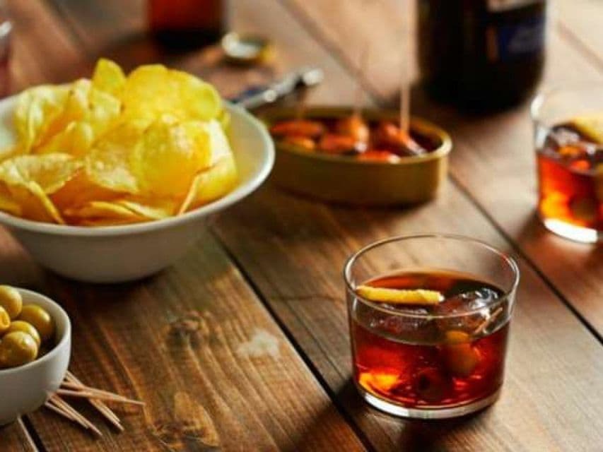An aperitif spread on a wooden table with cocktails, a bowl of potato chips, olives, and canned mussels.