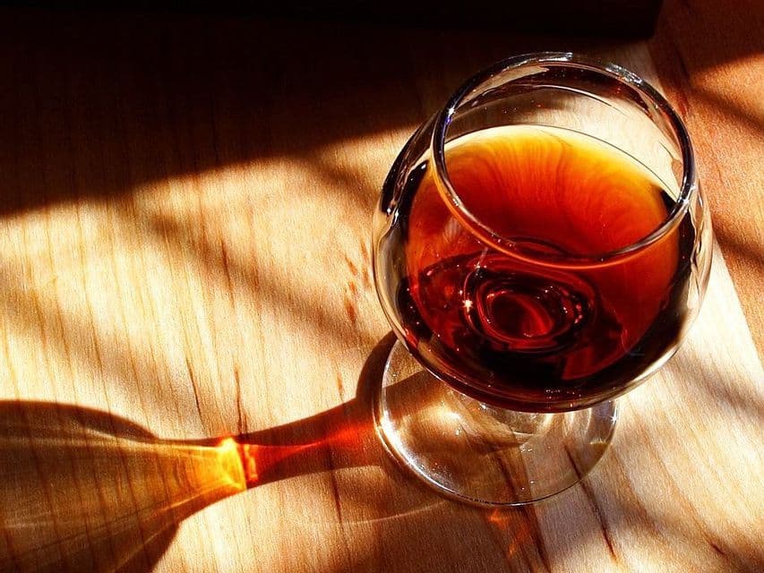 A snifter glass filled with dark liquor sits on a wooden surface, casting a long, colorful shadow from the sunlight.