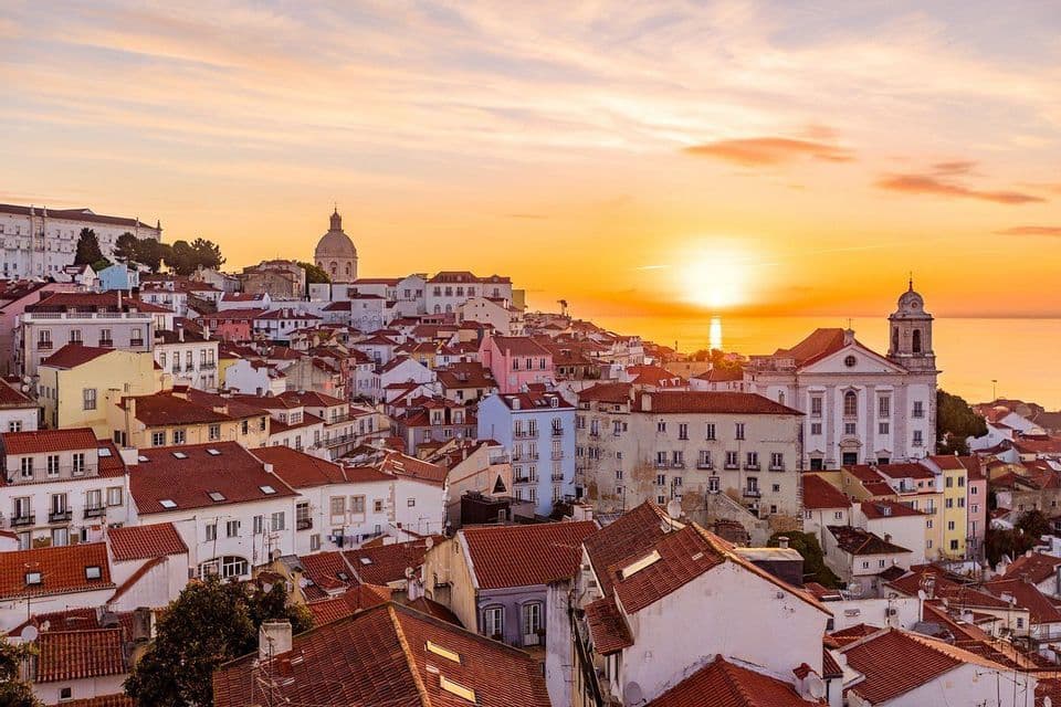 A cityscape of white buildings with red-tiled roofs on a hillside overlooking the water at sunset.