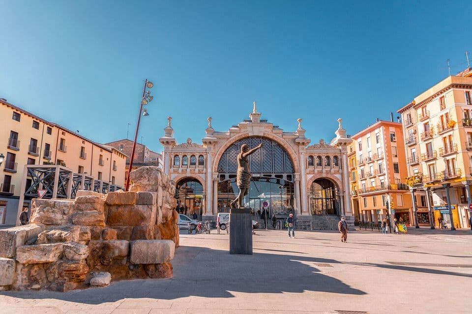 A sunny city plaza featuring a statue, ancient stone ruins, and an ornate historical building in the background under a clear blue sky.