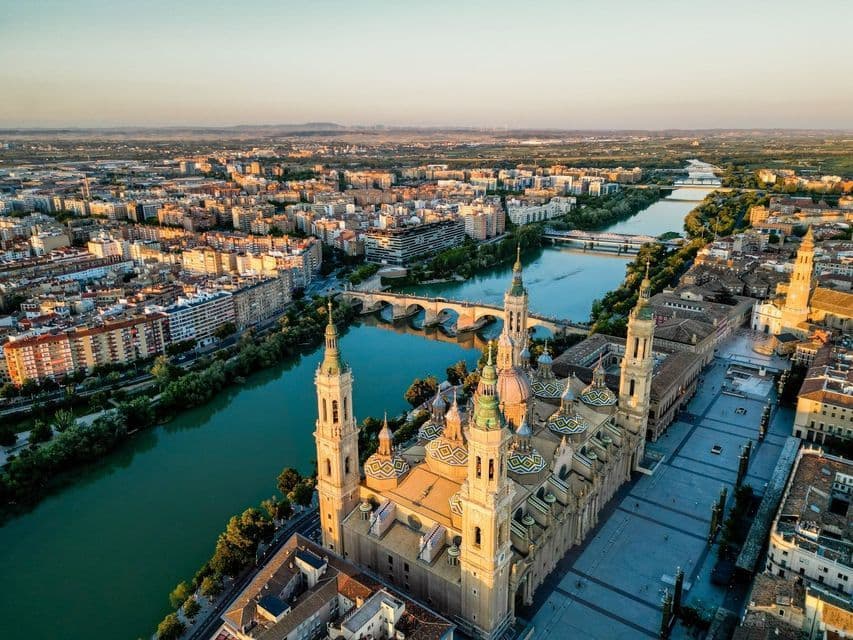 An aerial view of a large cathedral with ornate domes next to a river flowing through a city at sunrise.