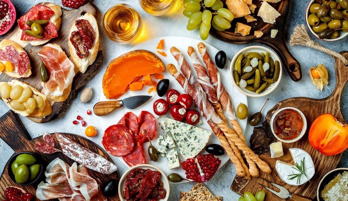 An overhead view of a large appetizer spread with assorted cheeses, charcuterie, fruits, olives, bread, and white wine.