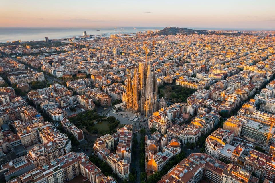 An aerial view of a large cathedral in the center of a dense cityscape at sunrise, with the sea in the background.