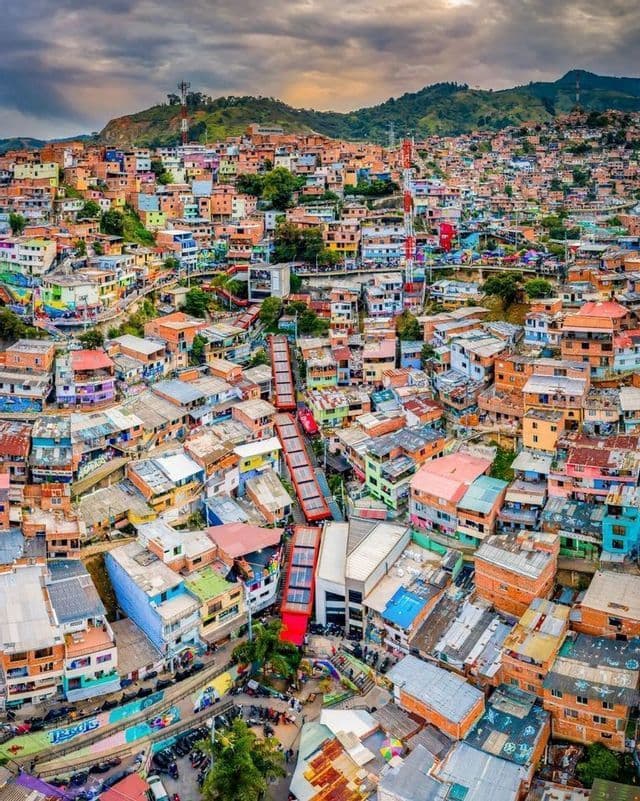 An aerial view of red escalators ascending through a densely packed, colorful hillside neighborhood under a cloudy sky.