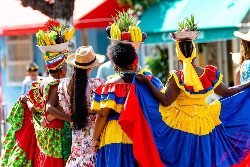 A group of women seen from behind, wearing colorful traditional dresses and carrying bowls of fruit on their heads.