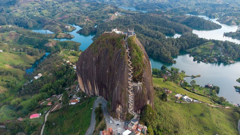 An aerial view of a giant monolith with a staircase built into its side, surrounded by a landscape of green hills and blue lakes.