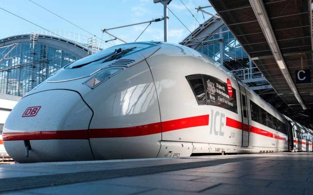 A low-angle shot of a white and red DB ICE high-speed train stopped at a station platform under a glass roof.