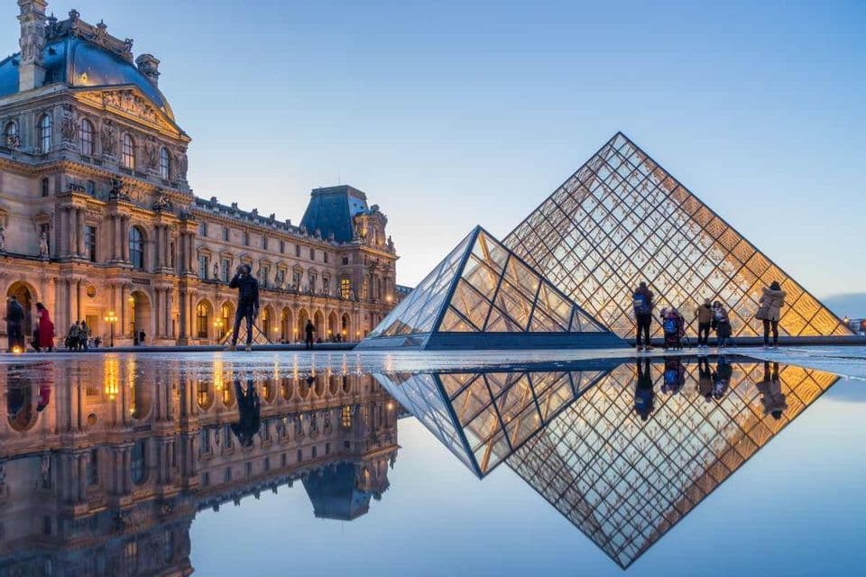 A glass pyramid and a classical palace are reflected in a large puddle on a courtyard at dusk, with people standing nearby.