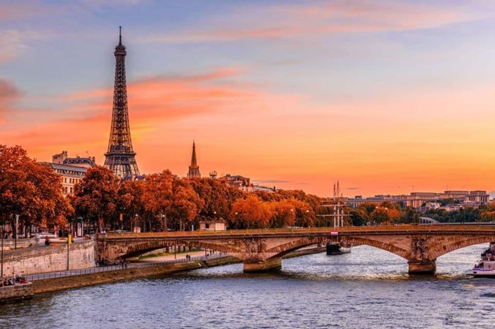 The Eiffel Tower and a bridge over the Seine river during a vibrant orange sunset, with autumn trees lining the riverbank.