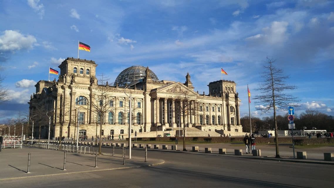A large, ornate government building with a glass dome and German flags flying on top, under a partly cloudy blue sky.