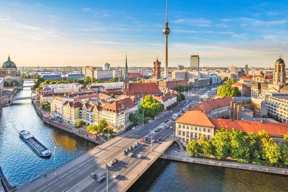 An aerial view of a city skyline with a prominent television tower, a river with a barge, and a bridge on a sunny day.