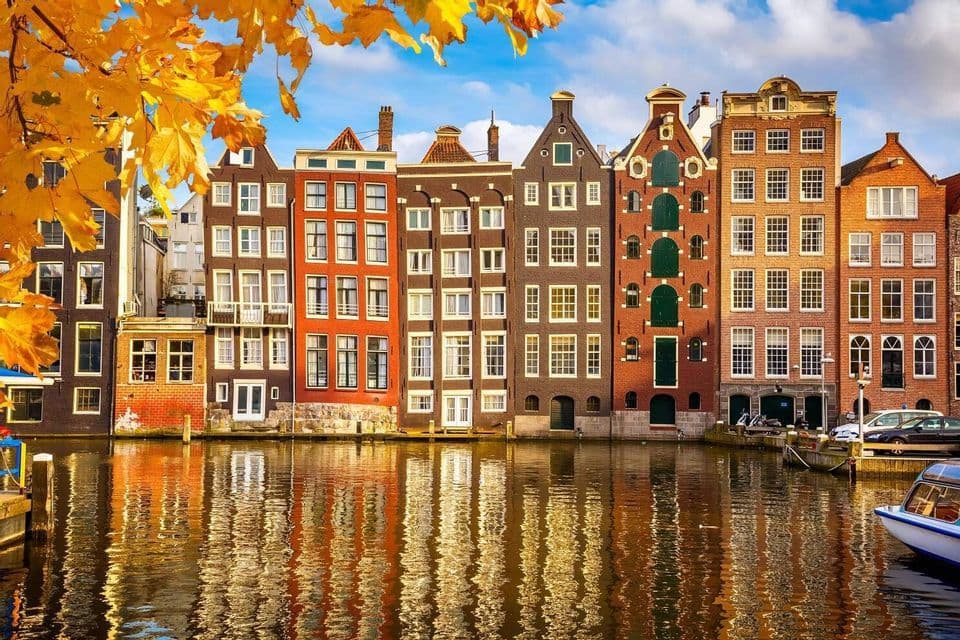 A row of colorful canal houses reflecting in the water, framed by yellow autumn leaves under a partly cloudy sky.