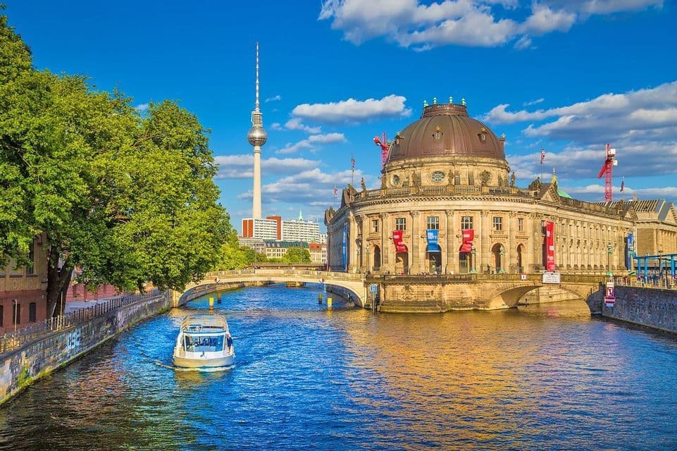 A tour boat sails on a river next to a grand domed building, with a tall television tower in the background under a blue sky.