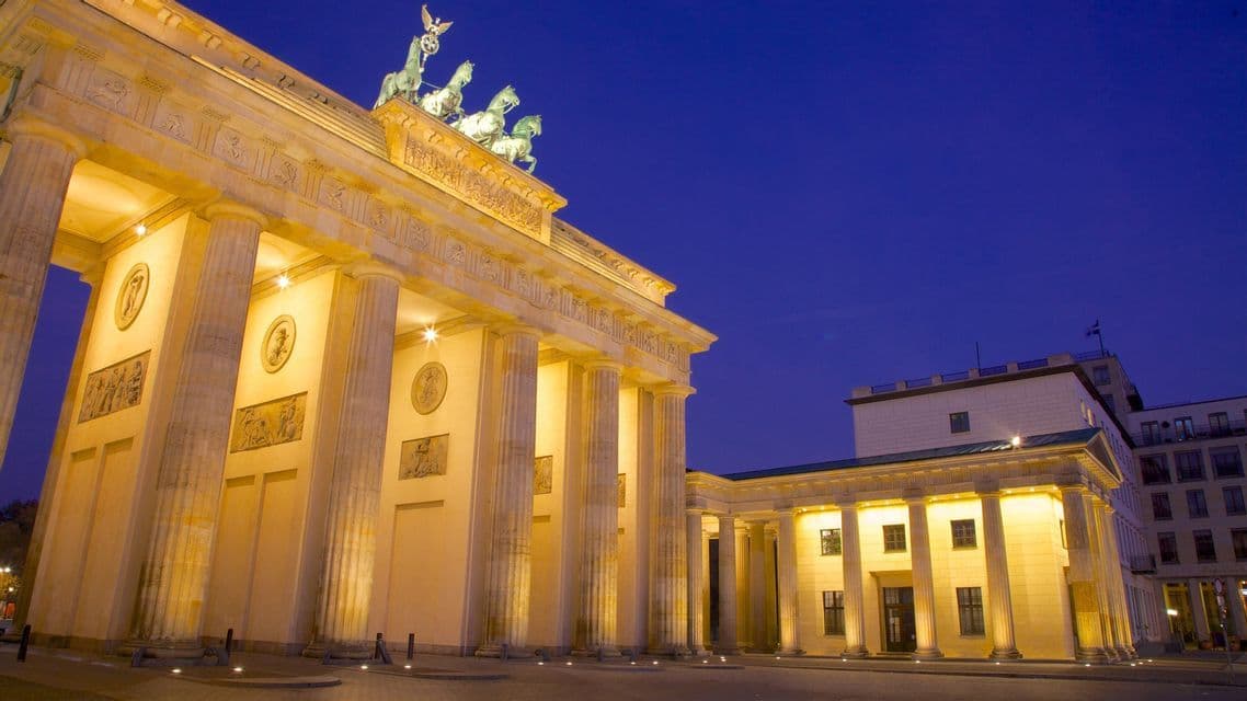 A large neoclassical arch topped with a quadriga statue is illuminated by warm lights against a deep blue twilight sky.