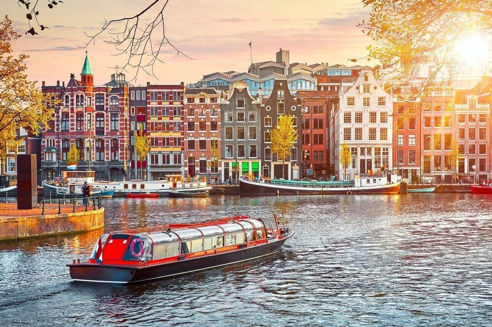 A red and black canal boat cruises past colorful, traditional gabled houses at sunset.